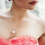 A woman wearing a vibrant red beaded dress and a delicate necklace with a square gemstone pendant.