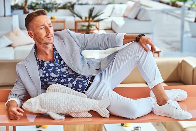 Man in a light gray blazer and white pants lounging on a bench with a decorative fish.