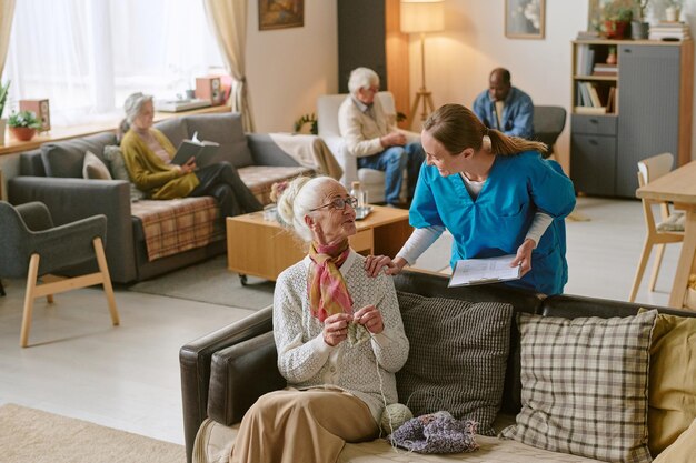 A caregiver assists an elderly woman in a cozy living room while other seniors relax nearby in a supportive community space.