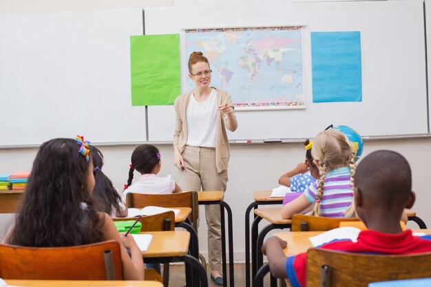 A teacher stands at the front of a classroom pointing toward students seated at desks, with a world map displayed on the board behind her.