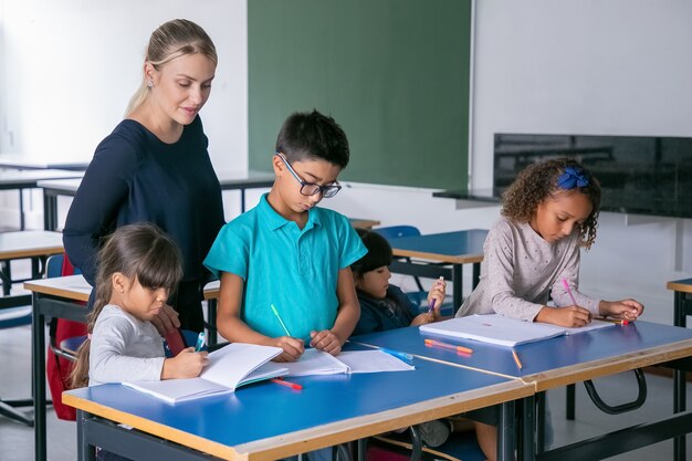 A teacher stands beside three children at a desk, helping them as they write in notebooks in a classroom.