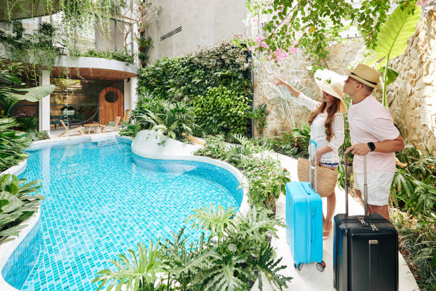 A vacationing couple holding suitcases near a pool in a tropical resort garden with vibrant plants and a relaxing atmosphere.