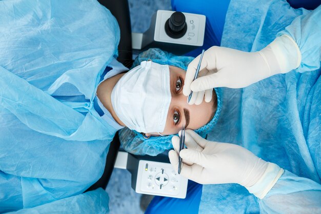 Patient lying on an operating table wearing a surgical mask and cap while a surgeon in gloves performs a cosmetic eye procedure using precision tools