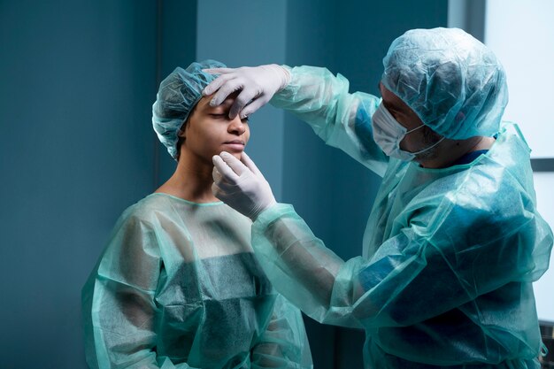 Surgeon in protective medical gear examining a patient’s face and forehead in a clinical setting, preparing for or assessing a cosmetic procedure.