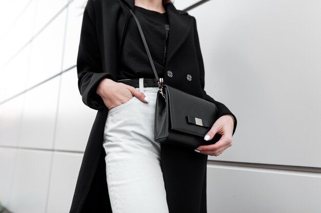 Woman in a black coat and white jeans carrying a crossbody handbag against a modern urban wall background.