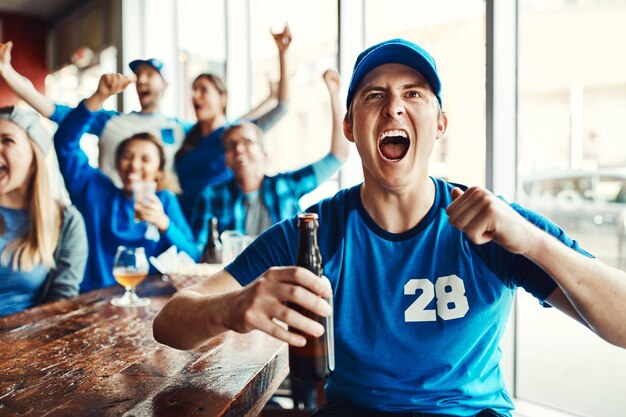 Group of sports fans in blue cheering in a bar, one man shouting and holding a beer bottle.