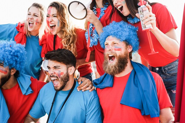 Excited fans in red and blue with face paint cheering loudly, one holding a megaphone and drinks.